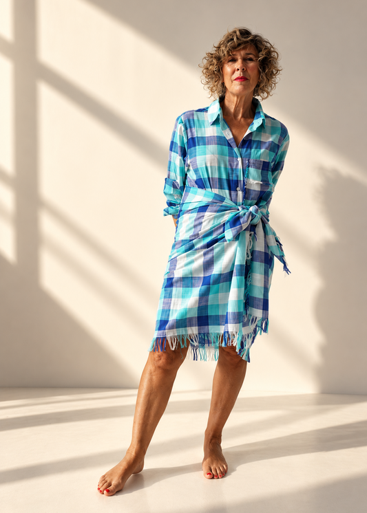 A woman modeling a blue, green, and white plaid wrap dress with a tie waist and fringed hem, standing against a warm lit studio background.