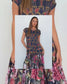 A woman modeling a tiered maxi dress with a navy blue base featuring a gold chain-link pattern on the bodice and a colorful floral print on the skirt.