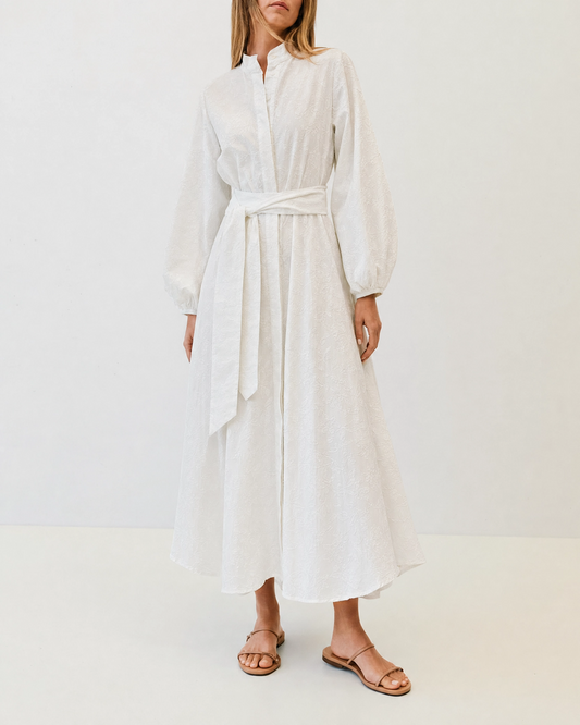 A woman wearing a white long-sleeve midi shirt dress with a tie waist belt, paired with brown leather flat sandals, standing against a plain white studio background.
